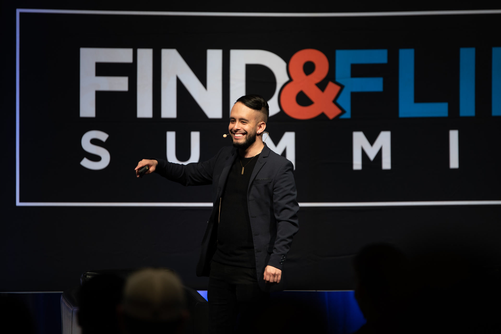 Ulyses on stage at Find & Flip Summit, navy blazer, headset mic, clicker, Find & Flip backdrop.