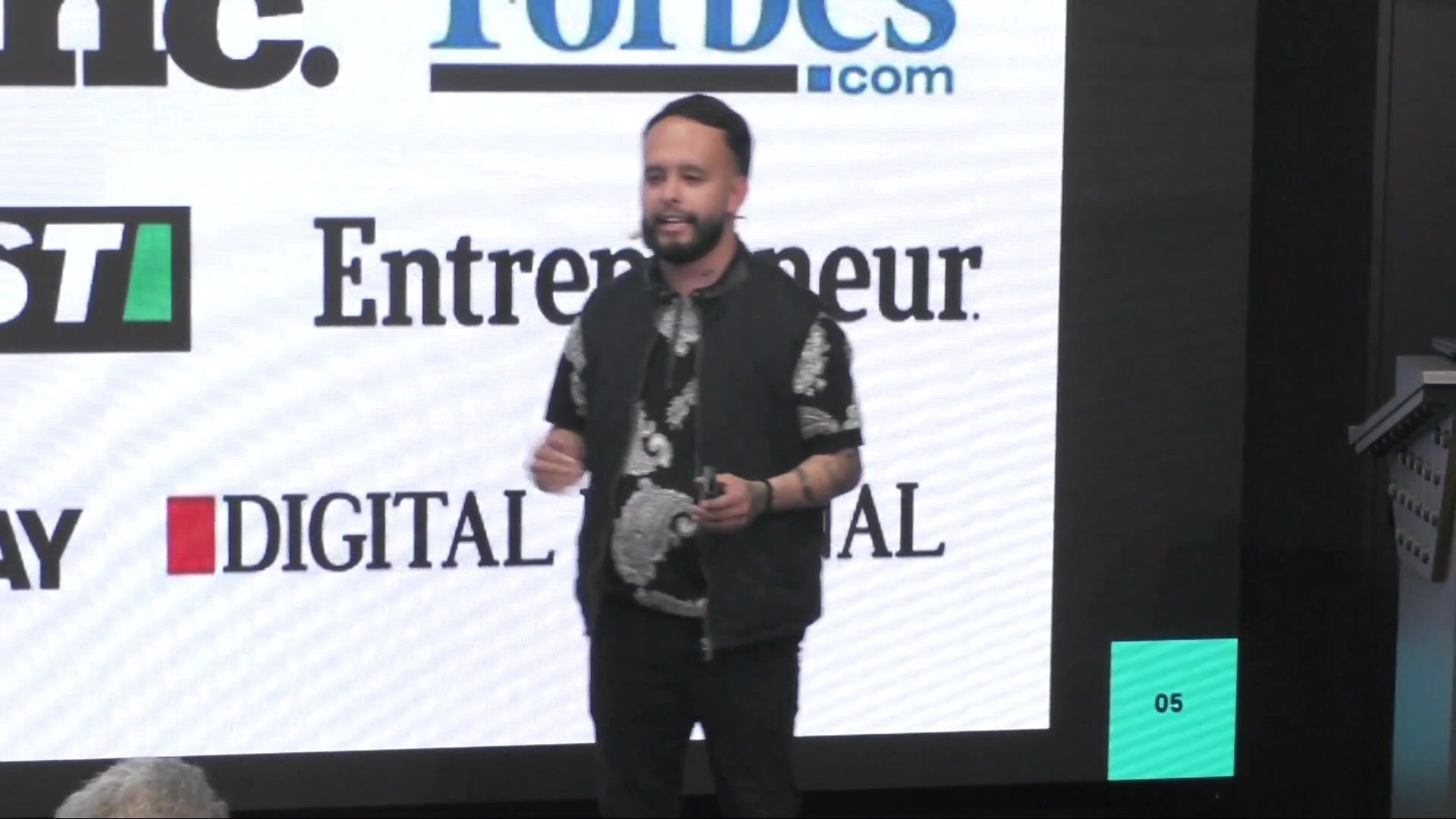 Speaker in a paisley shirt and black vest in front of a Forbes.com / Entrepreneur / Inc. / Digital Journal logo wall.