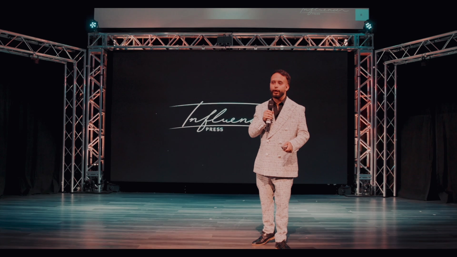 Speaker in a light textured suit on a trussed stage; screen reads Influencer PRESS.