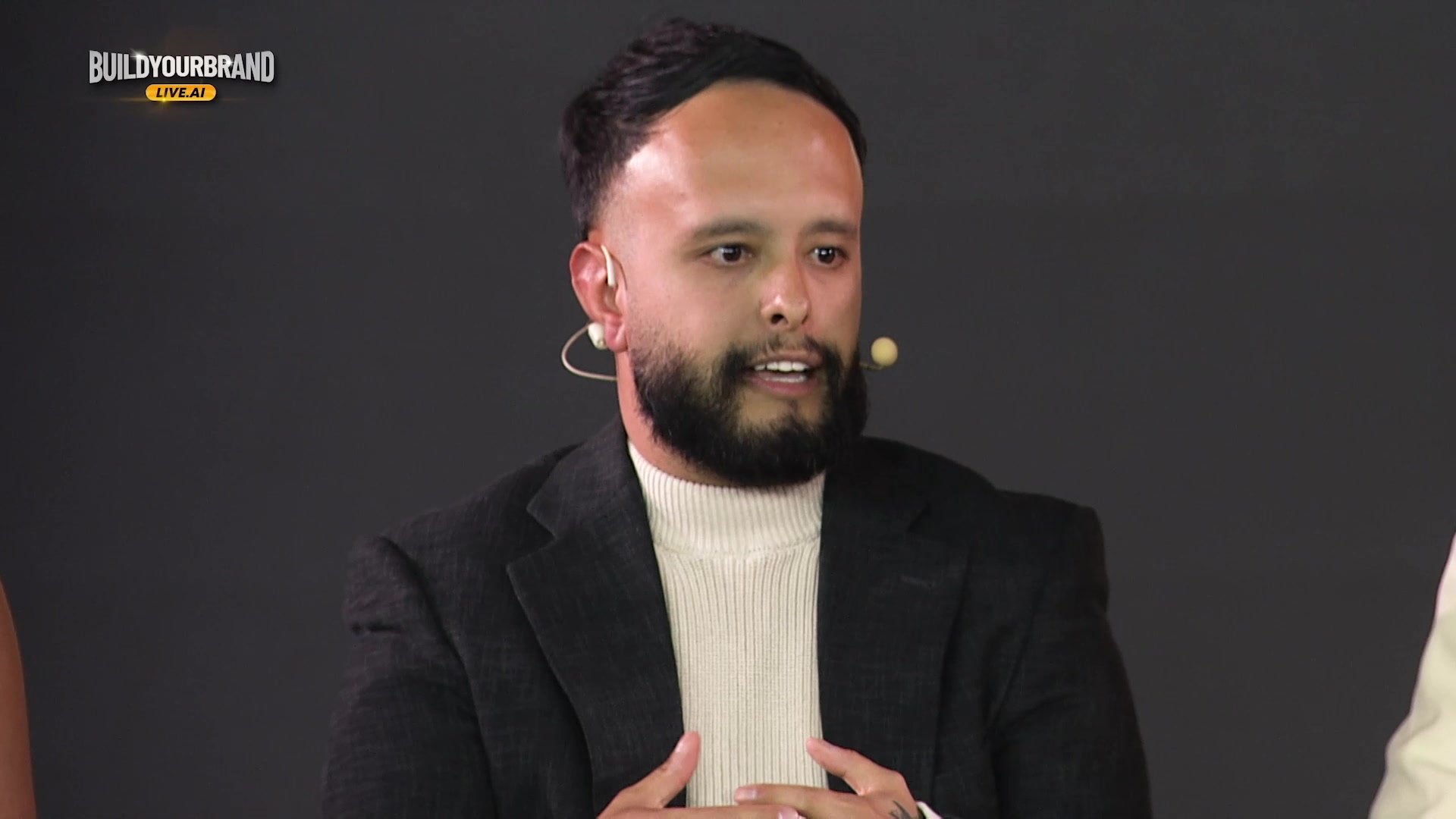 Close-up, charcoal blazer and cream turtleneck, headset mic; partial Build Your Brand Live.ai mark.