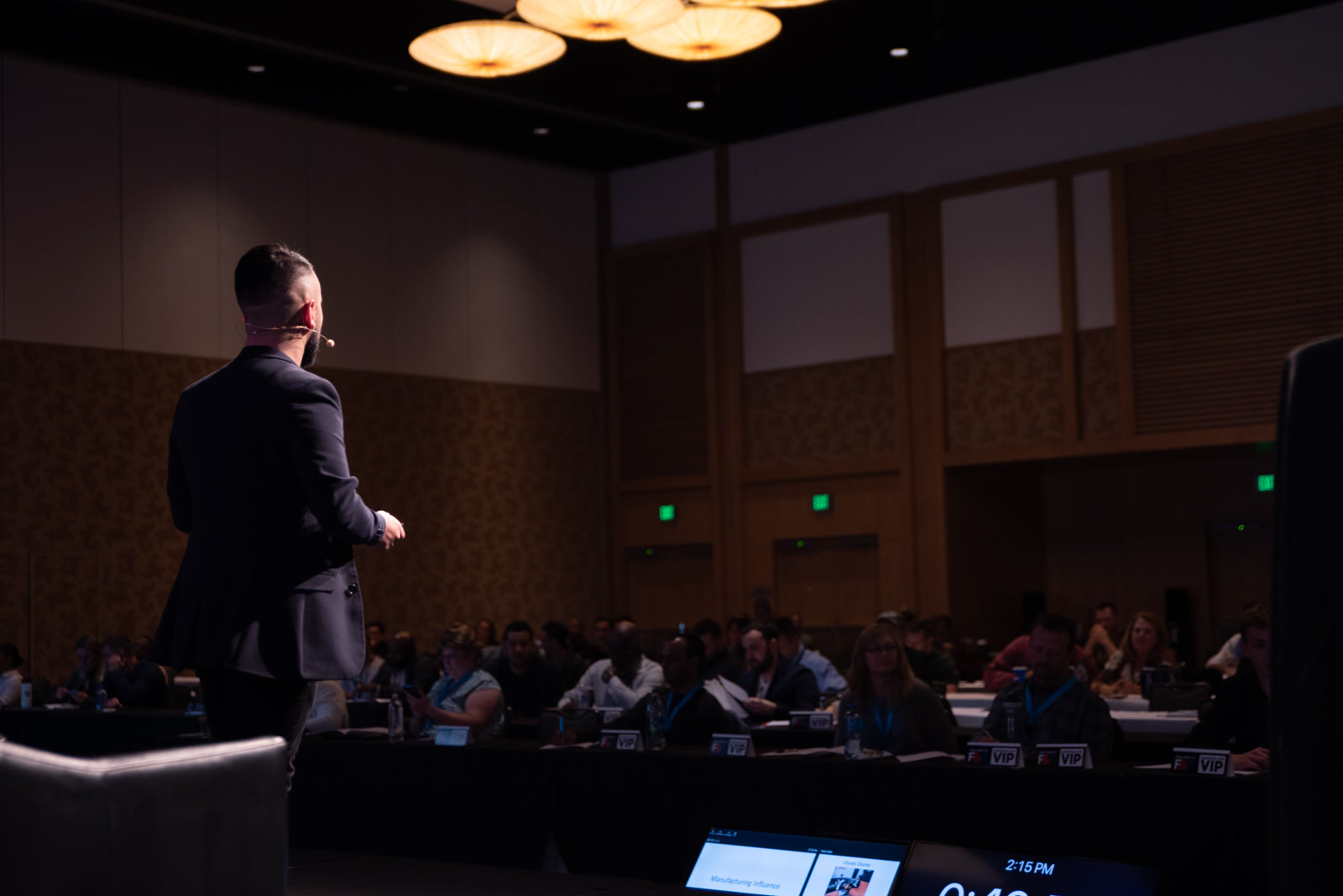 Speaker on stage facing a seated audience, headset mic on, confidence monitors in the foreground.